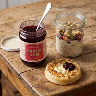Jar of berry jam, jar of muesli, crumpet with jam and cream on a wooden table