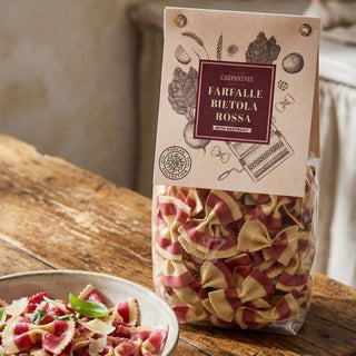 Bag of Carpinini's Farfalle Bietola Rossa pasta with a bowl of pasta on a wooden table.
