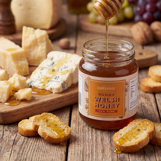Jar of Carpaninis Runny Welsh Honey with honey being drizzled over it, surrounded by cheese and bread on a wooden table.