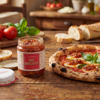 Jar of Spicy Bruschetta sauce on a wooden table with a pizza and bread slices.