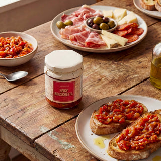 A jar of spicy bruschetta on a wooden table with bread and cheese