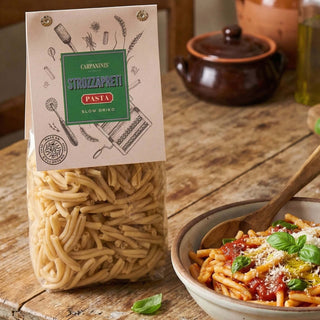 Bag of Carpaninis Strozzapreti pasta on a wooden table with a bowl of pasta in the background.