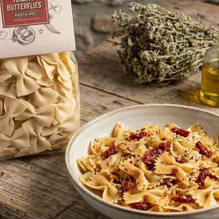 Pasta dish with sun-dried tomatoes next to a package of bow tie pasta on a wooden surface.